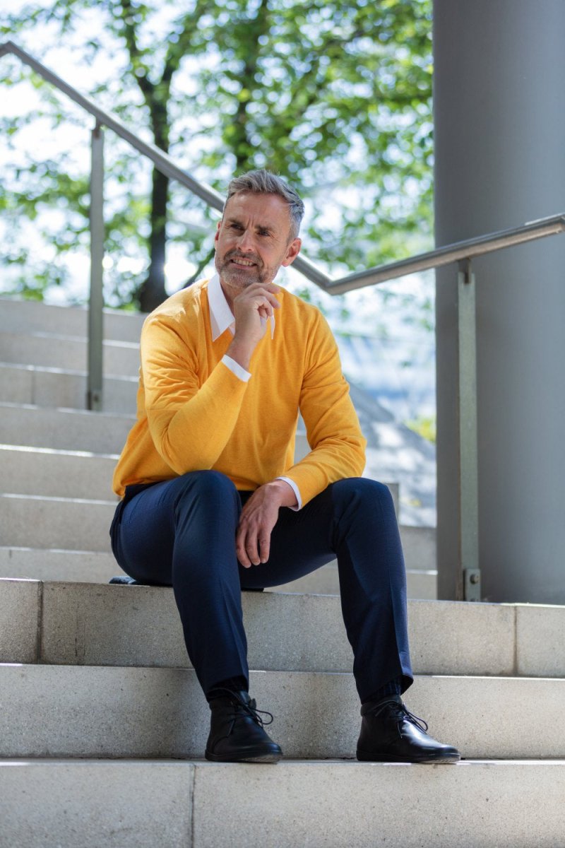 Ein Mann mit grauem Haar und Bart sitzt nachdenklich auf einer Außentreppe. Er trägt einen leuchtend gelben Pullover über einem weißen Hemd und dunkelblauen Hosen. Seine schwarzen Schuhe sind poliert. Im Hintergrund sind ein Metallgeländer, eine Säule und grüne Bäume im klaren Sonnenlicht zu sehen.