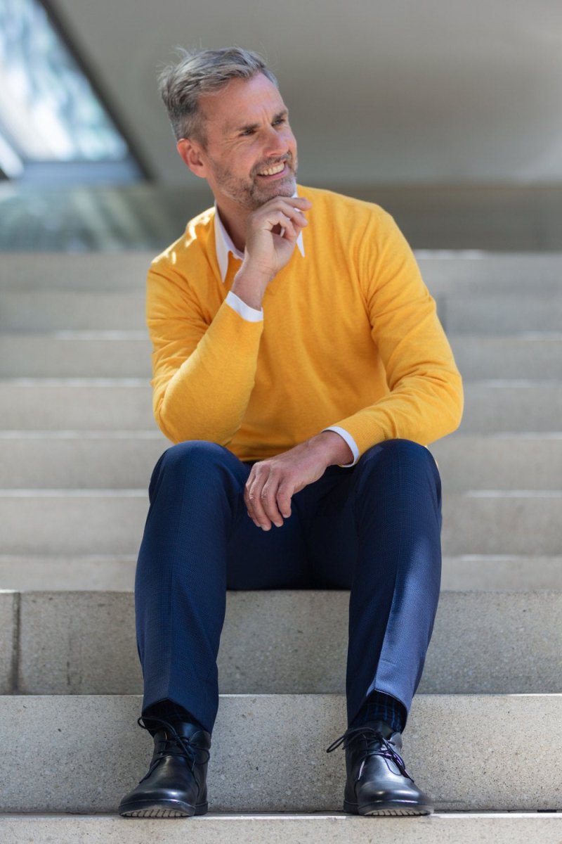 Ein Mann mit grauem Haar und Bart sitzt auf einer Außentreppe. Er trägt einen leuchtend gelben Pullover über einem weißen Hemd, blaue Hosen und schwarze Schuhe. Er blickt zur Seite, lächelt und stützt sein Kinn auf seine Hand. Der Hintergrund ist unscharf und lässt einen sonnigen Tag vermuten.