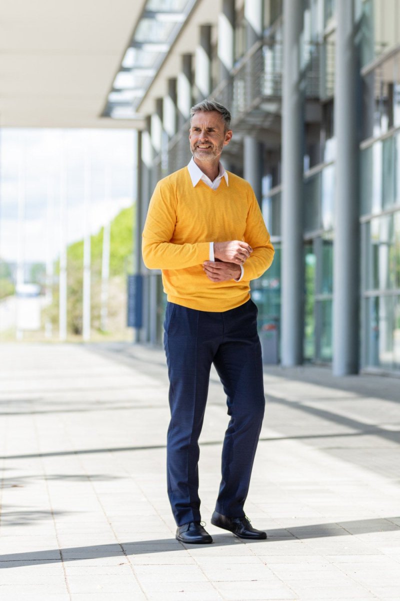 Ein Mann mit grauem Haar und Bart steht selbstbewusst im Freien neben einem modernen Gebäude. Er trägt einen leuchtend gelben Pullover über einem weißen Hemd mit Kragen und dunkle Hosen sowie schwarze Schuhe. Die Sonne wirft Schatten auf den Bürgersteig und im Hintergrund sind grüne Bäume zu sehen.
