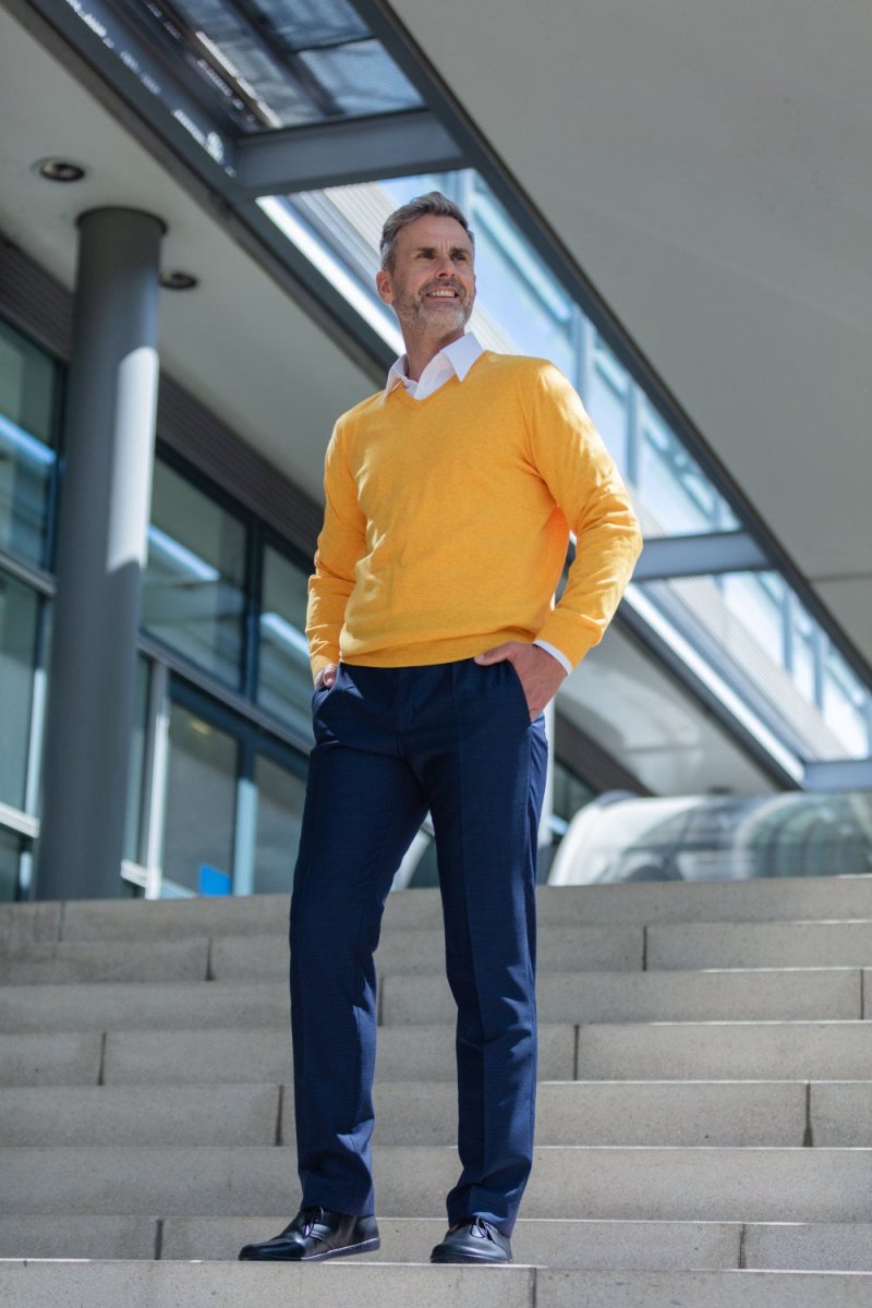 Ein Mann steht selbstbewusst auf den Stufen vor einem modernen Gebäude. Er trägt einen leuchtend gelben Pullover über einem weißen Hemd und dunkelblauen Hosen. Seine Hände sind in den Taschen und er lächelt leicht. Im Hintergrund sind Glasfenster und Stahlträger unter einem bewölkten Himmel zu sehen.