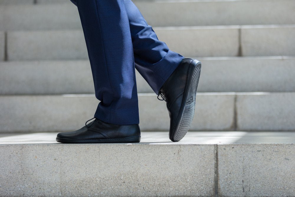 Eine Person in marineblauer Anzughose und schwarzen Lederschuhen steigt Betonstufen hinauf. Der Fokus liegt auf den Unterschenkeln und Füßen, wodurch die Bewegung des Aufsteigens eingefangen wird. Die Schuhe haben einen dezenten Glanz und die Stufen sind gut beleuchtet, was das saubere und moderne Gefühl der Umgebung hervorhebt.